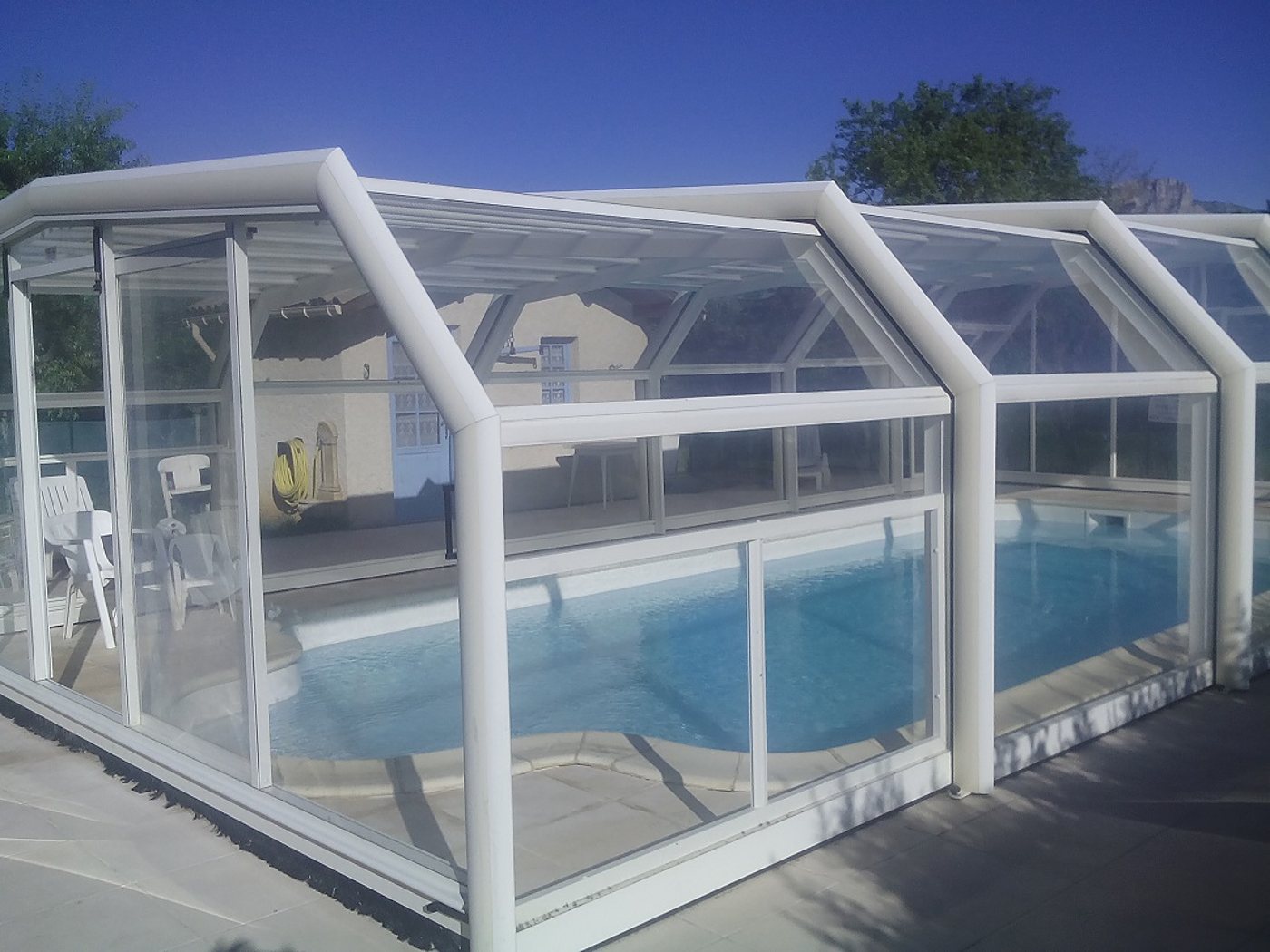 Un abri de piscine haut à structure blanche et parois transparentes recouvre un bassin d'eau bleue, avec une maison et des chaises de jardin visibles à l'arrière.