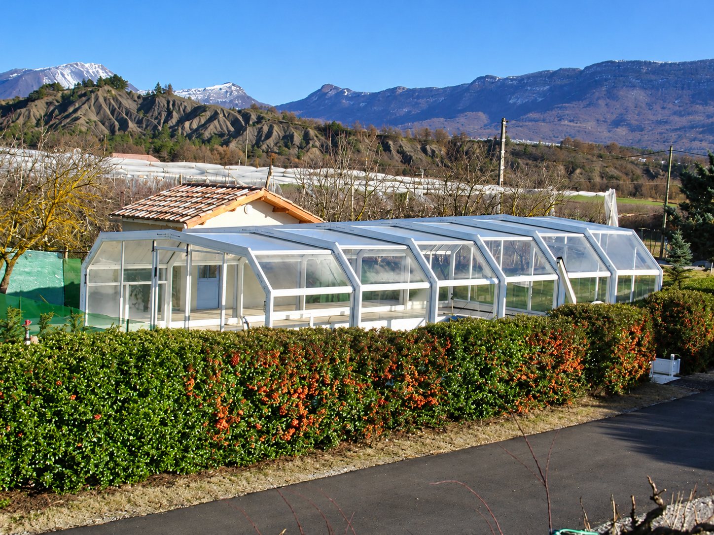 Un abri de piscine télescopique blanc et vitré est installé à côté d'une maison au toit de tuiles, derrière une haie fleurie, avec des montagnes au loin.