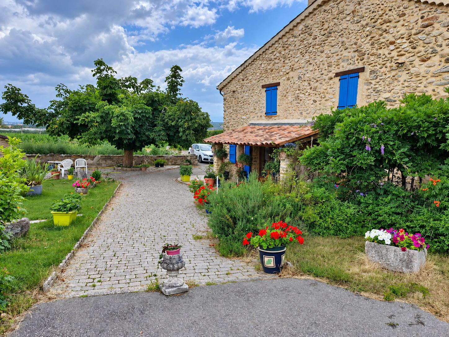 Une allée de pavés bordée de pots de fleurs et de massifs mène à une maison en pierres aux volets bleus, sous un ciel nuageux.