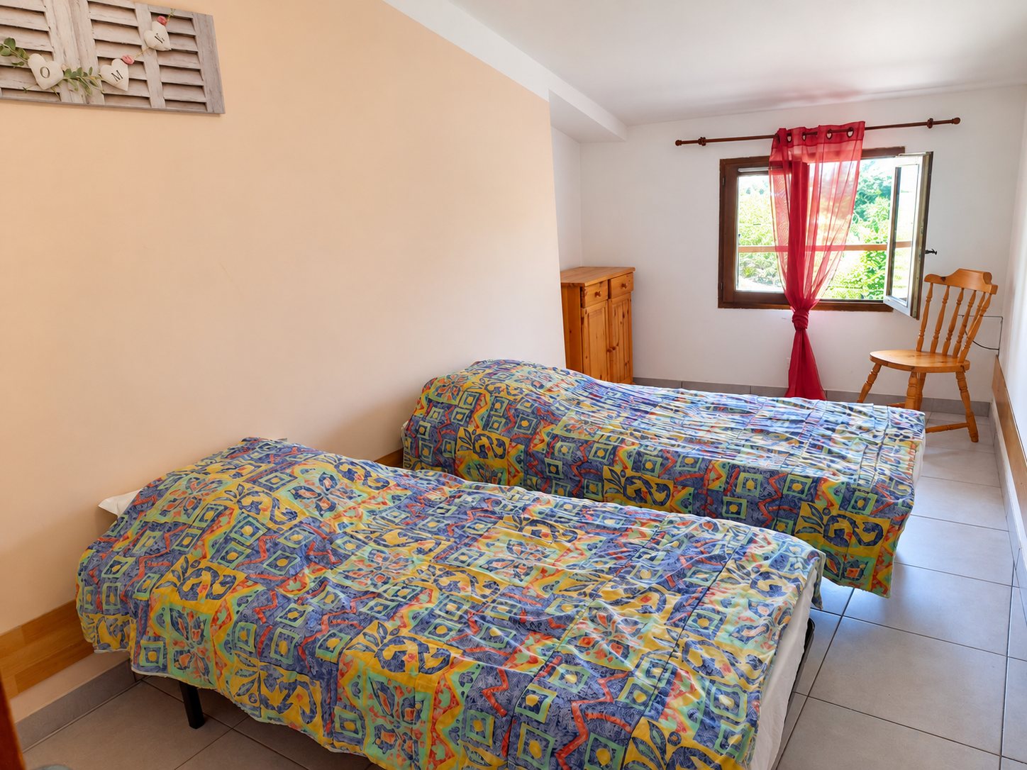 Chambre au carrelage clair avec deux lits simples couverts de plaids colorés, une commode en bois, une chaise et une fenêtre ouverte avec rideau rouge.