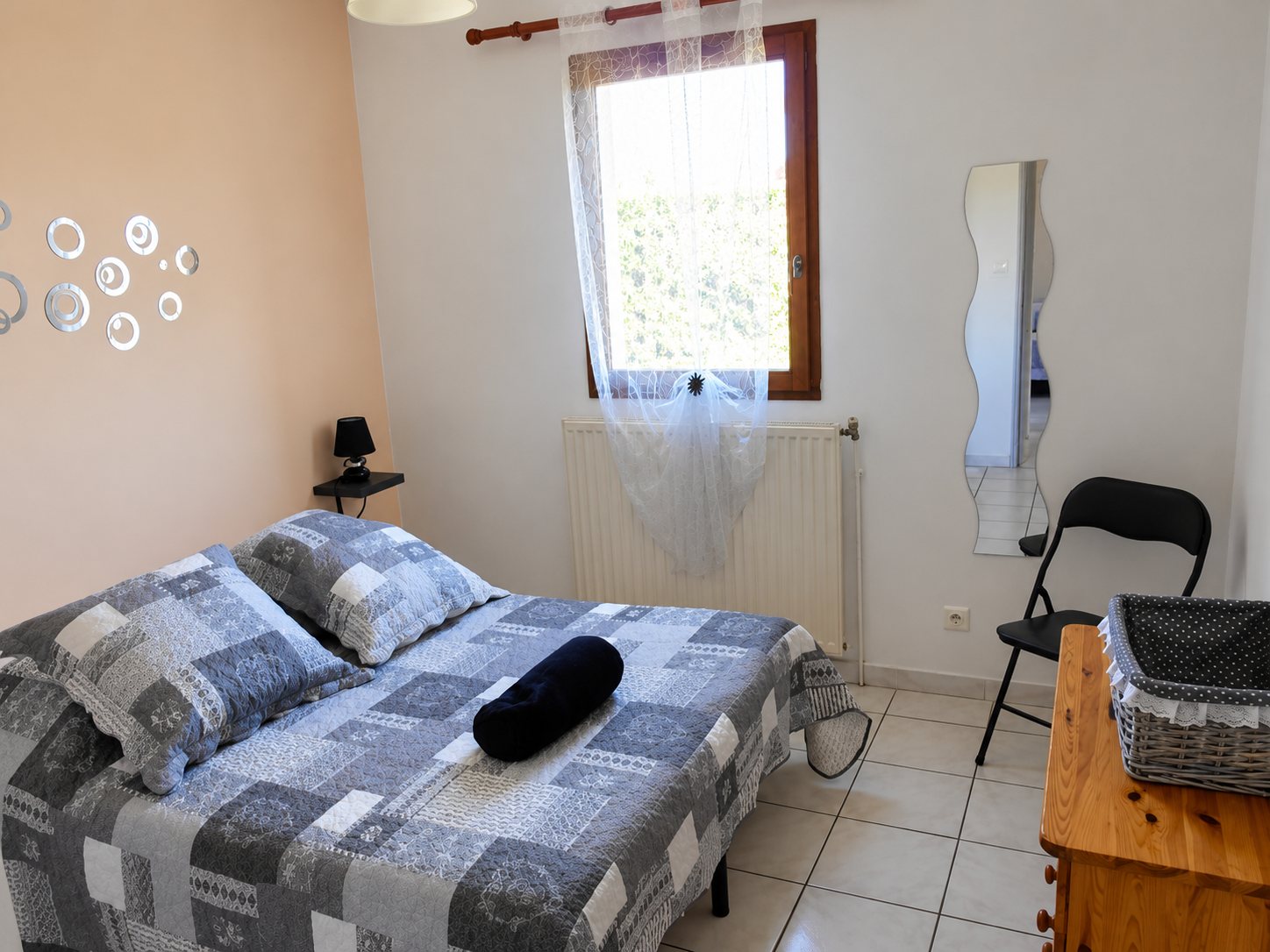 Une chambre claire avec un lit double recouvert d'un patchwork bleu et gris, une fenêtre voilée, une chaise noire pliante et une commode en bois portant un panier.
