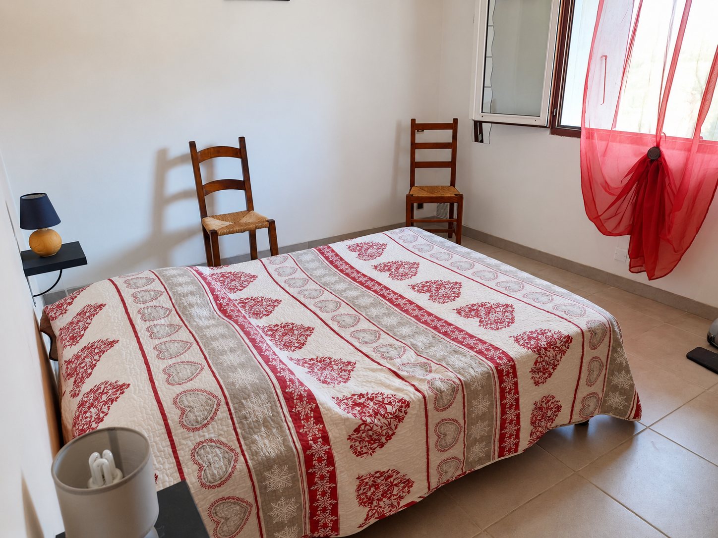 Chambre claire avec un lit double recouvert d'un couvre-lit à motifs de cœurs rouges, deux chaises en paille, une lampe de chevet et un rideau rouge à la fenêtre.
