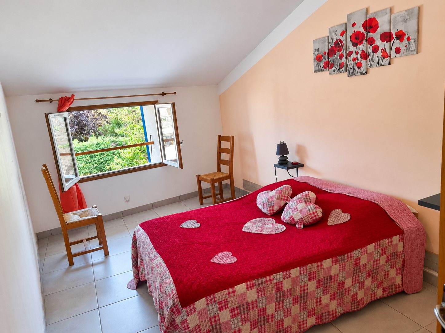 Chambre sous les combles avec un grand lit recouvert d'un couvre-lit rouge orné de cœurs en tissu, deux chaises en bois, une fenêtre ouverte sur la verdure et un tableau de coquelicots au mur.