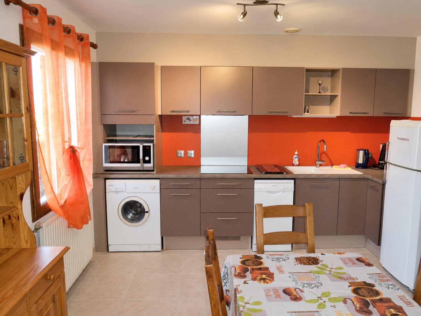 Cuisine aménagée aux meubles beige-brun et à la crédence orange, avec lave-linge, réfrigérateur blanc et table recouverte d'une nappe motif café.