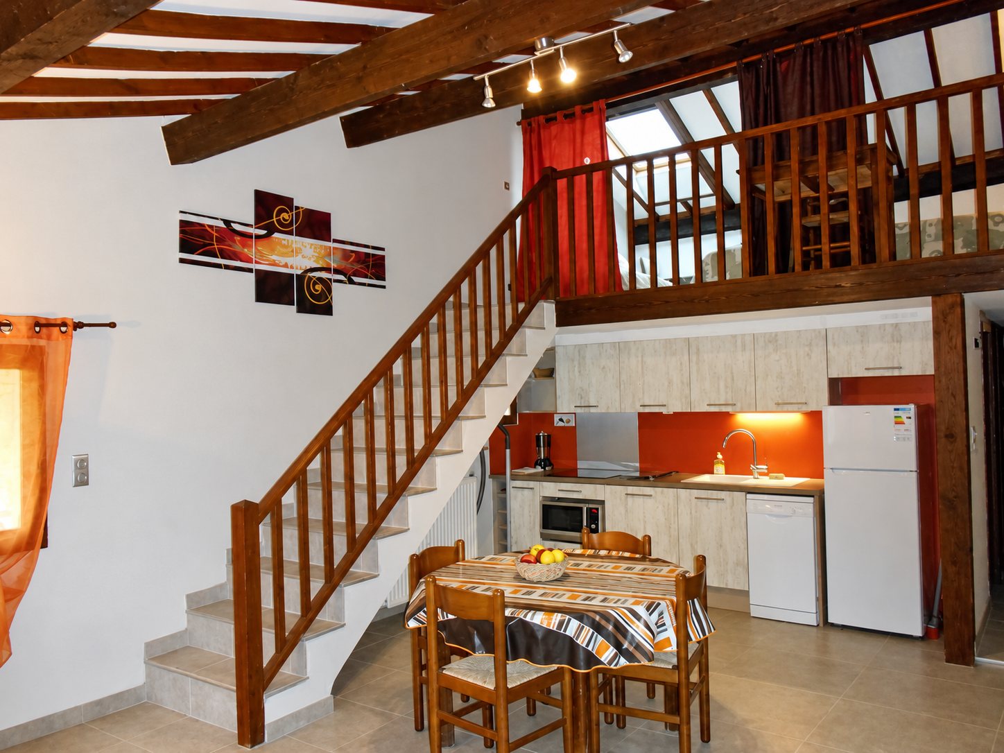 Pièce de vie avec cuisine équipée orange et blanche, table ronde avec corbeille de fruits, escalier en bois montant à une mezzanine sous des poutres apparentes.