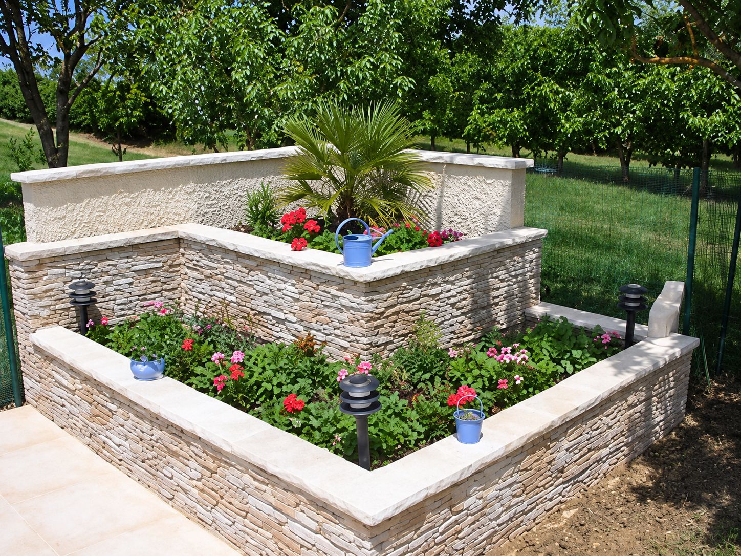Grande jardinière d'angle à plusieurs niveaux en pierre naturelle, plantée de fleurs rouges et d'un palmier, dans un jardin verdoyant.