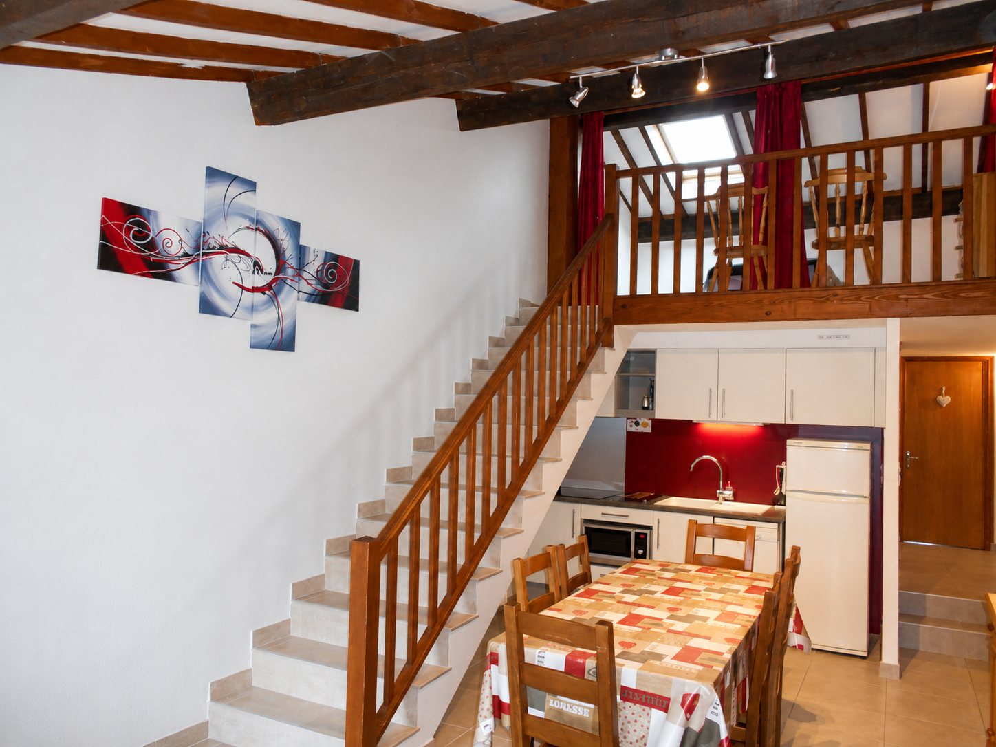 Intérieur d'un gîte avec une cuisine équipée rouge et blanche, une table dressée, un escalier en bois menant à une mezzanine sous des poutres apparentes.