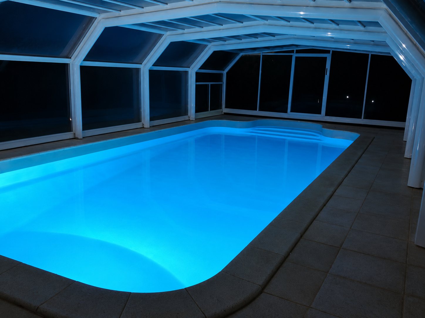 Une piscine rectangulaire illuminée en bleu dans l'obscurité, protégée par un abri télescopique blanc et entourée de dalles sombres.