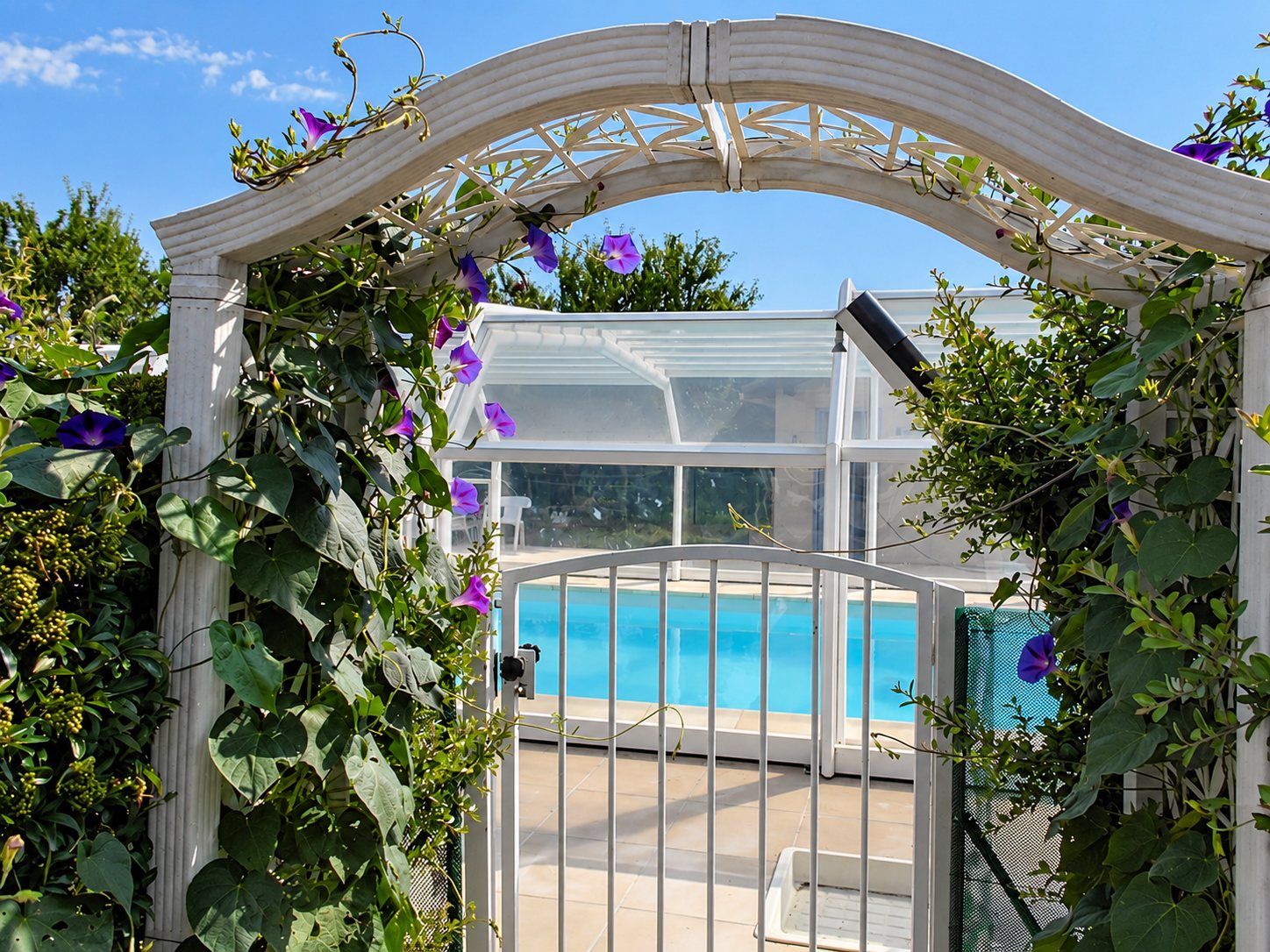 Une arche blanche couverte de liserons violets encadre un portillon qui ouvre sur une piscine protégée par un abri vitré.