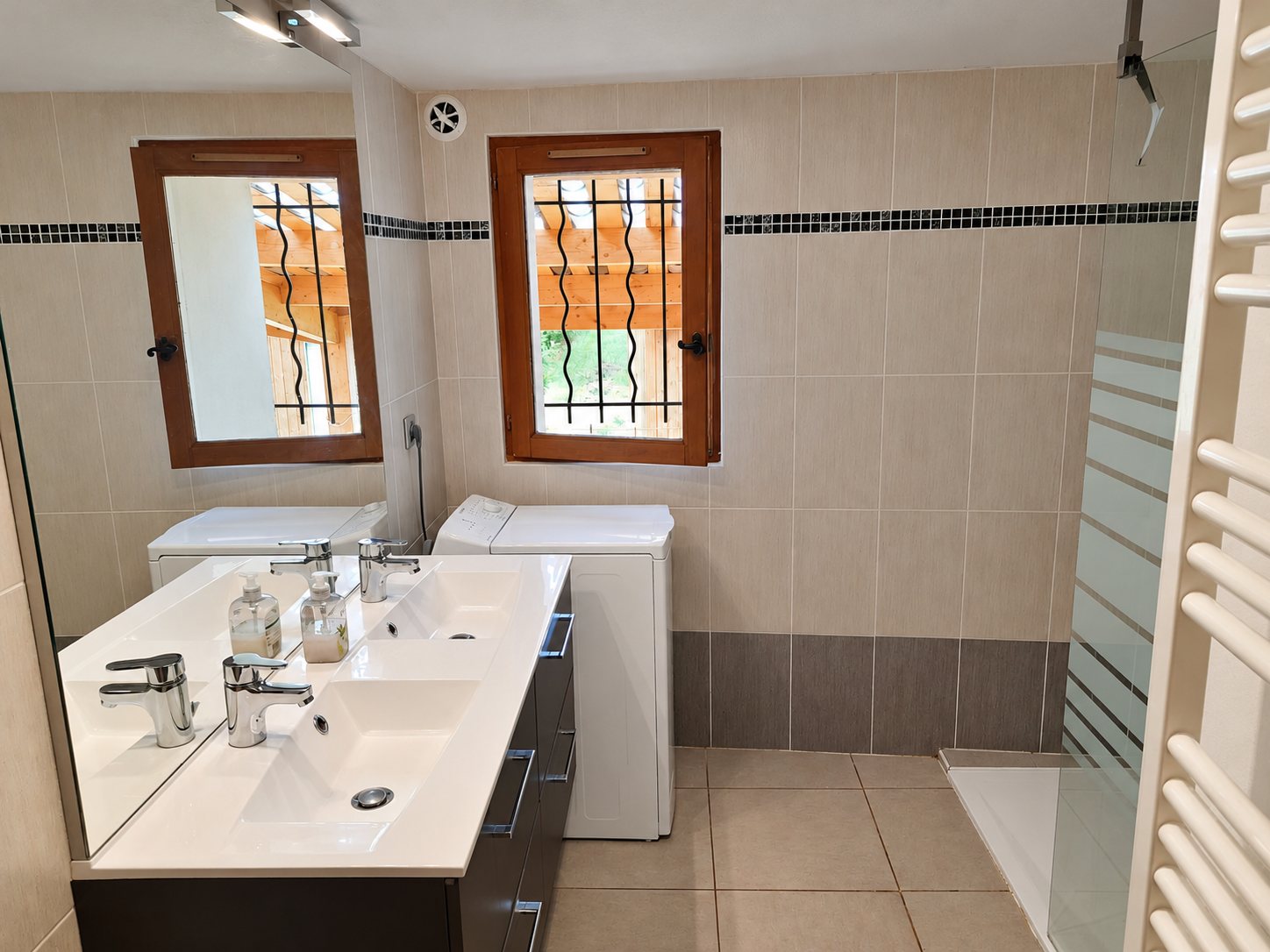 Salle de bain carrelée beige avec meuble à double vasque et grand miroir, lave-linge sous une fenêtre à barreaux, douche vitrée et sèche-serviettes blanc.