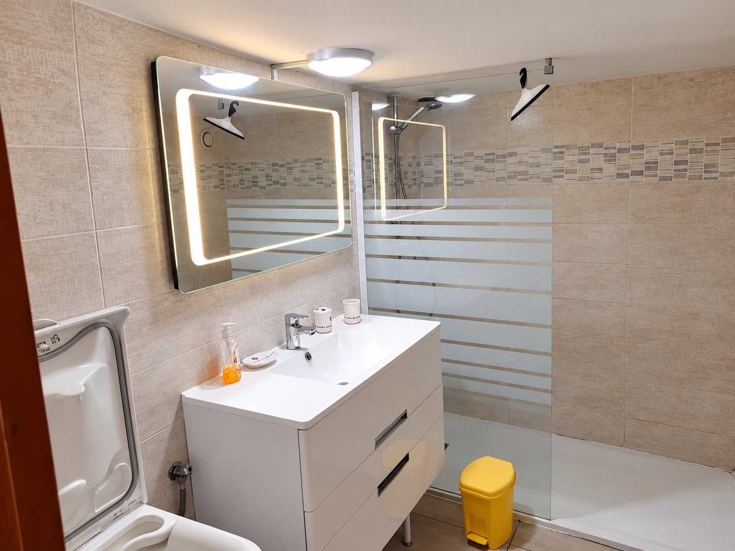 Salle de bain carrelée en beige avec un meuble vasque blanc, un miroir lumineux, une douche à l'italienne avec paroi en verre dépoli et une petite poubelle jaune.